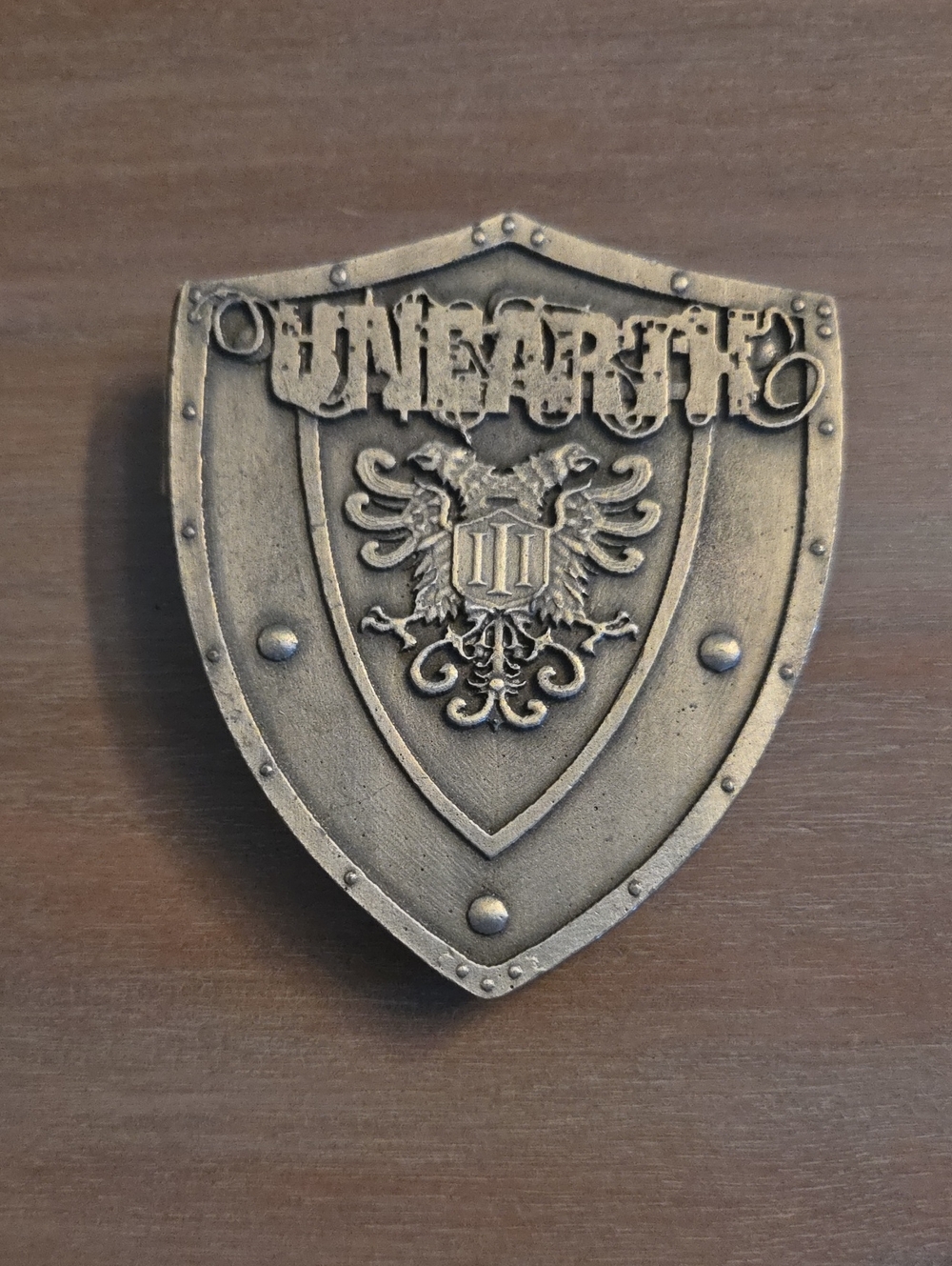 official Unearth shield logo belt buckle, featuring a distressed, antique bronze